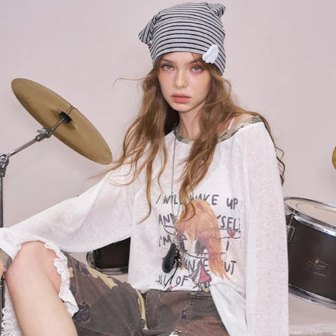 Person sitting on a drum set wearing a white oversized shirt with graphics and camouflage pants, with 'ELF SACK' branding.